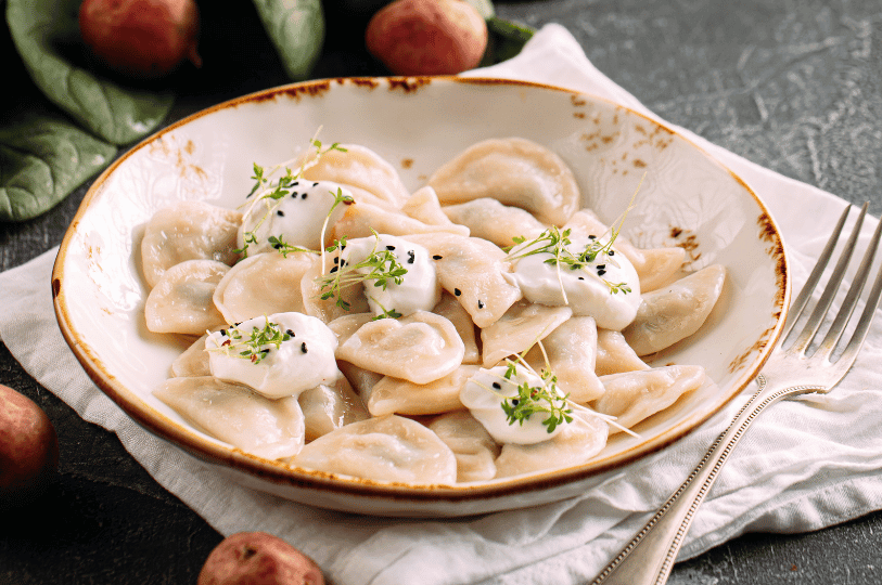 A bowl of boiled pierogi served with creamy white sauce and fresh micro-herbs, presented in a rustic ceramic bowl on a white linen napkin.
