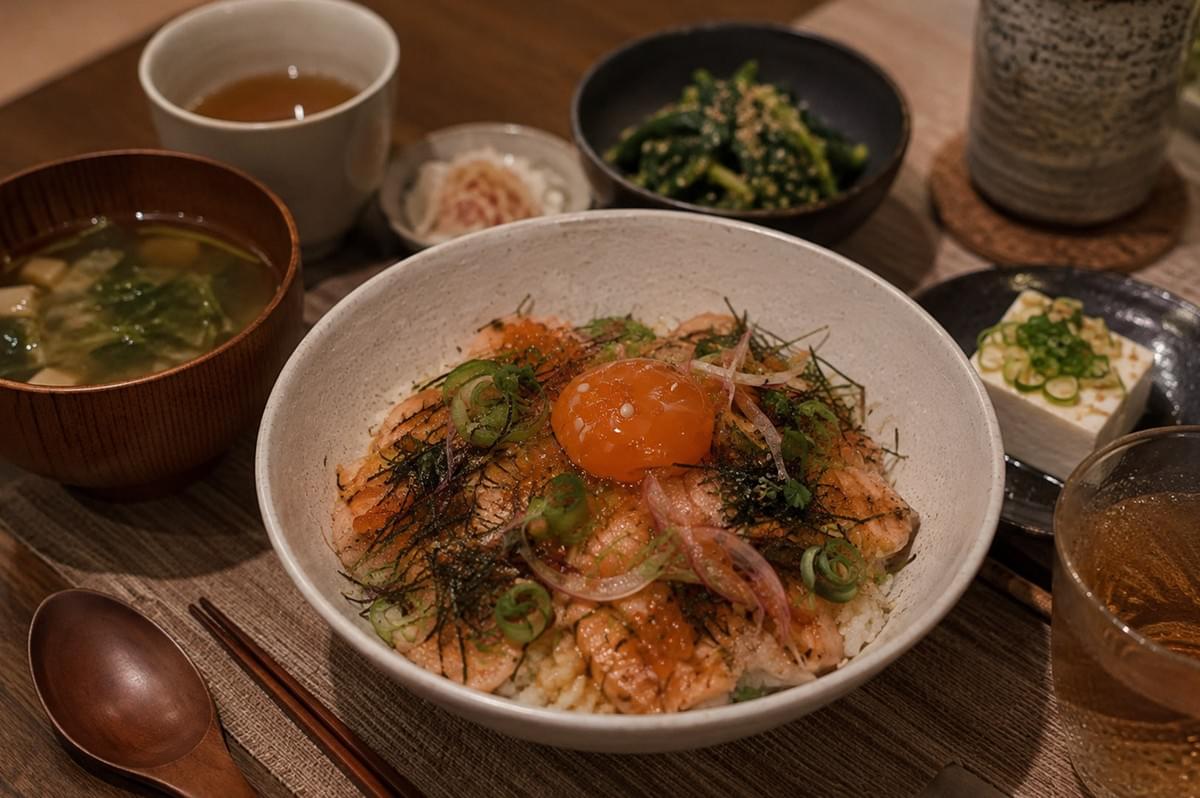 A warm, cozy dining shot presents a complete Japanese set meal featuring the aburi salmon chirashi bowl at the center on a woven placemat. Surrounding the main bowl are side dishes including a wooden bowl of hot miso soup with tofu and greens, a small dish of cold tofu topped with scallions, a bowl of sesame-dressed spinach side dish (gomaae), a cup of hot tea, and a glass of iced tea alongside wooden chopsticks and a spoon.