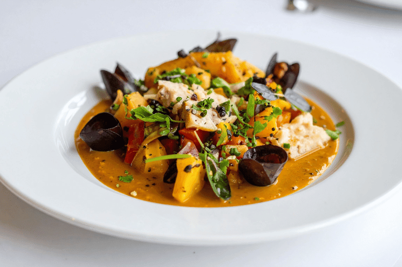 A white bowl containing a modern interpretation of Provencal Bouillabaisse or a regional seafood stew, featuring chunks of white fish, mussels, and golden potatoes in a thick, saffron-hued broth garnished with fresh microgreens.
