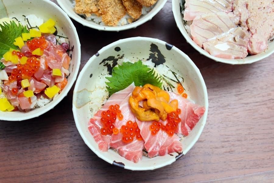 Four ceramic bowls of Japanese rice dishes are arranged on a wooden table, with the central bowl featuring sliced raw tuna, bright orange sea urchin, and shiny salmon roe over white rice. Surrounding bowls display other seafood preparations, including diced raw fish with yellow pickled radish, crispy fried cutlets, and seared fish slices.