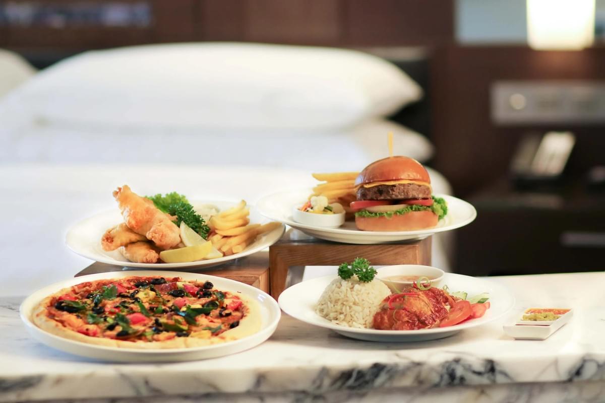 A room service spread features four plates of food, including a pizza, a cheeseburger with fries, fish and chips, and chicken served with rice. The dishes are arranged on a marble counter in front of a softly blurred hotel bed with crisp white linens.