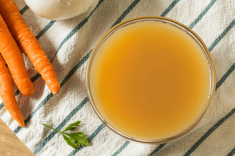 An overhead view of a glass bowl filled with opaque, savory chicken stock, surrounded by raw carrots and parsley on a striped kitchen towel.