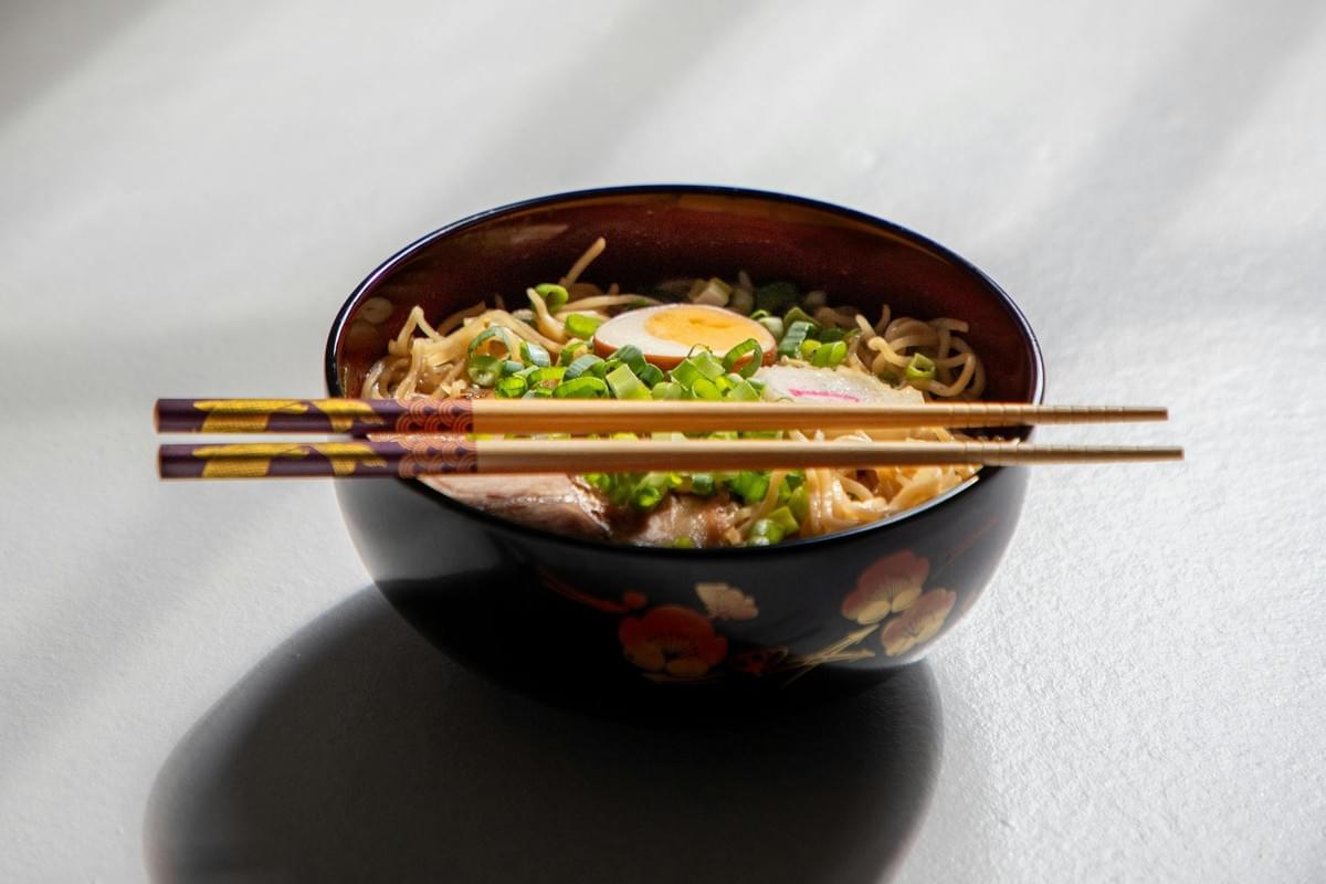 A decorative black bowl filled with ramen noodles is topped with chopped green onions, a slice of fish cake, and a halved soft-boiled egg. Resting horizontally across the rim is a pair of patterned chopsticks, illuminated by bright light that casts a distinct shadow on the plain surface below.