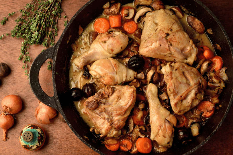 Overhead view of a black Dutch oven filled with Coq au Vin, surrounded by raw ingredients like onions and bundles of fresh herbs.