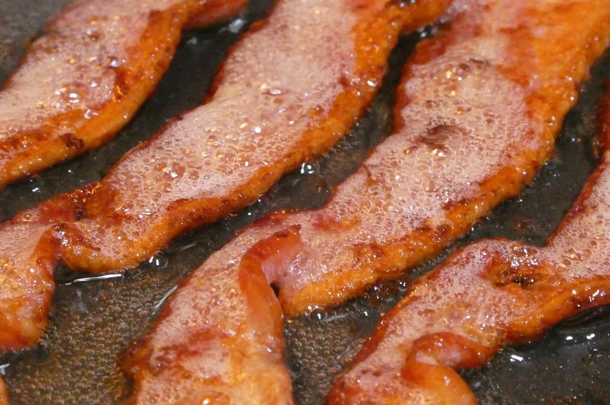 Bacon sizzling in a frying pan on a stove, part of a recipe using salt pork, drippings, and aromatics.