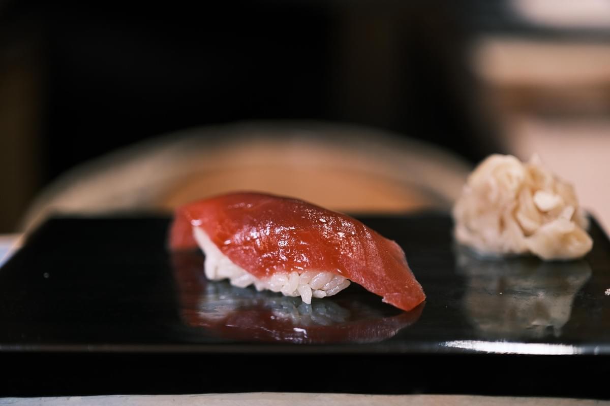 A piece of lean tuna nigiri sits on a sleek black lacquered plate, accompanied by a small mound of pickled ginger. The shot uses a shallow depth of field, making the glossy texture of the fish stand out against a softly blurred background.