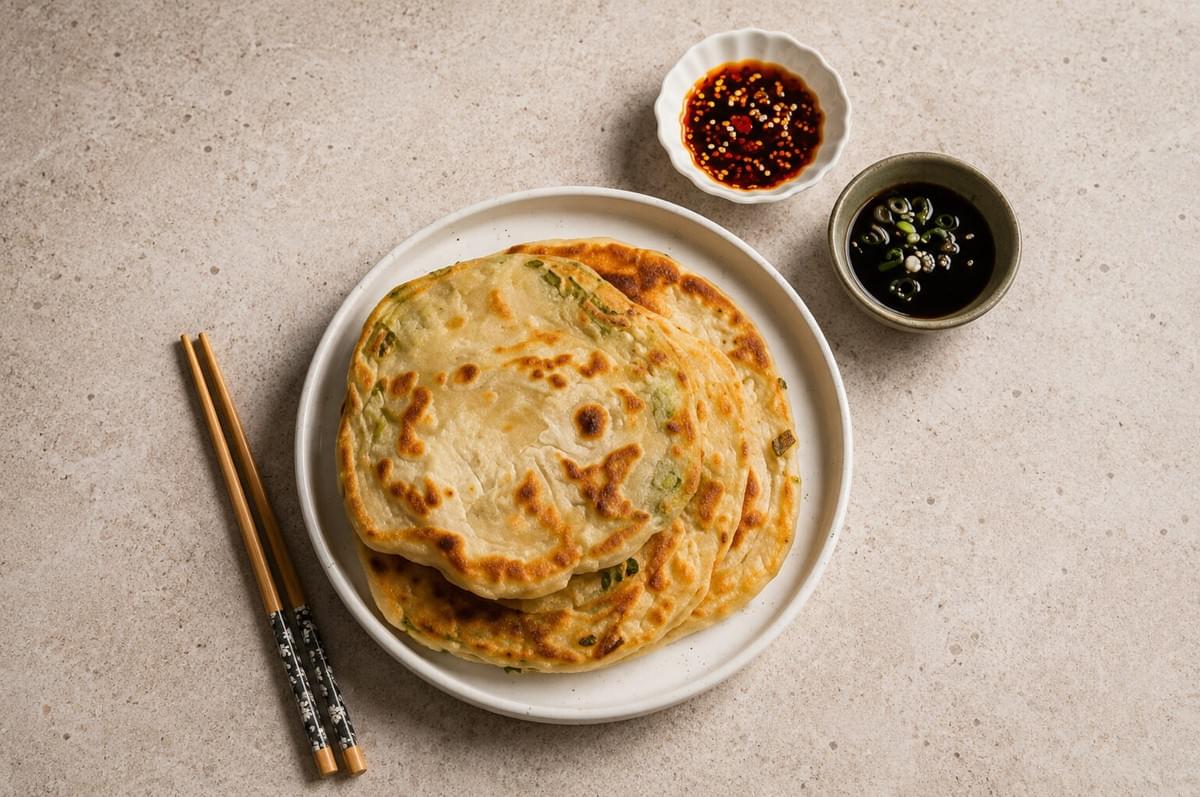 Pan-fried scallion pancakes on a white plate next to chopsticks and dipping sauces.