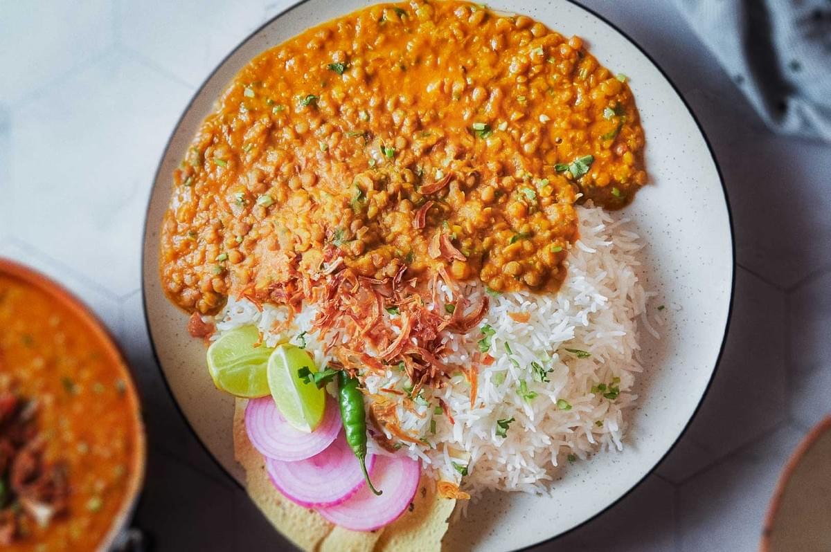 Indian dal curry with basmati rice topped with fried onions, garnished with cilantro, served with papadum, red onion slices, lime wedges, and green chili.