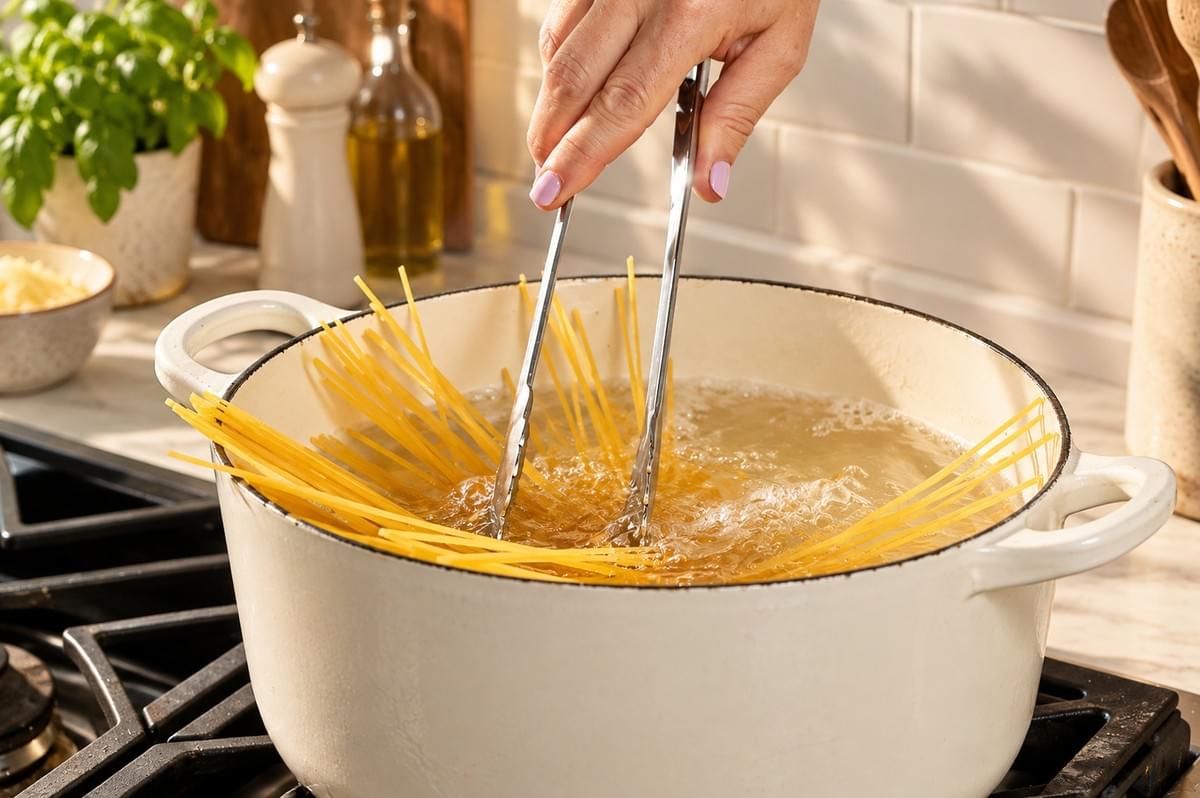Metal tongs pushing uncooked spaghetti noodles into a pot of boiling water on a stovetop.