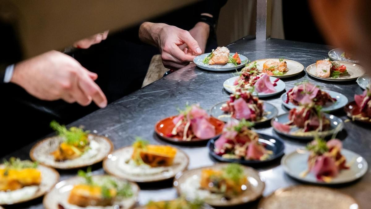 This image is taken from a slightly obscured, high angle, showing two chefs' hands plating a large number of appetizers or small dishes on a dark kitchen counter.
