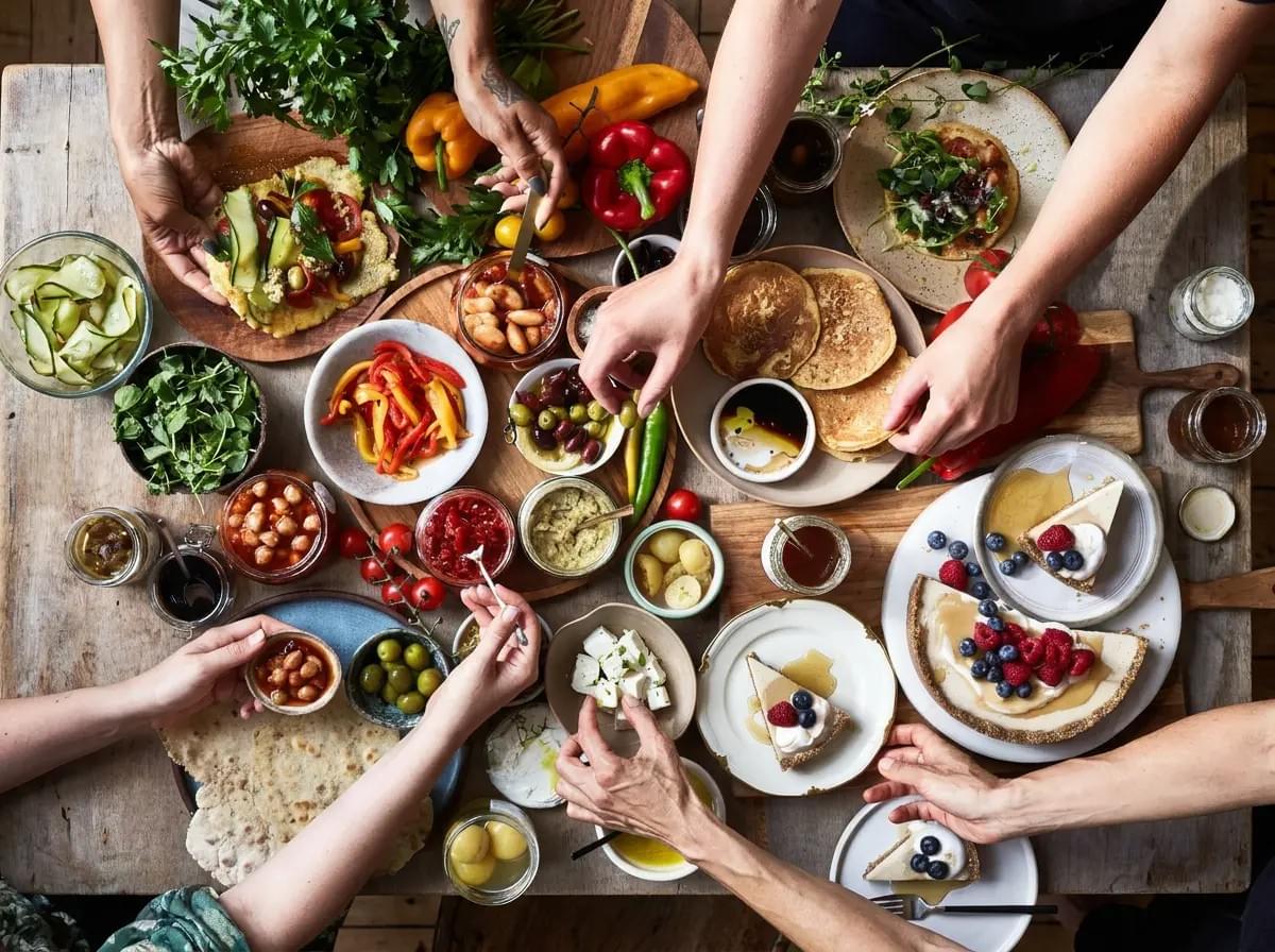 An overhead view of hands reaching into a busy rustic table spread with Mediterranean-style brunch dishes, including flatbreads, pancakes, feta cheese, olives, fresh vegetables, jams, and berry-topped tarts.
