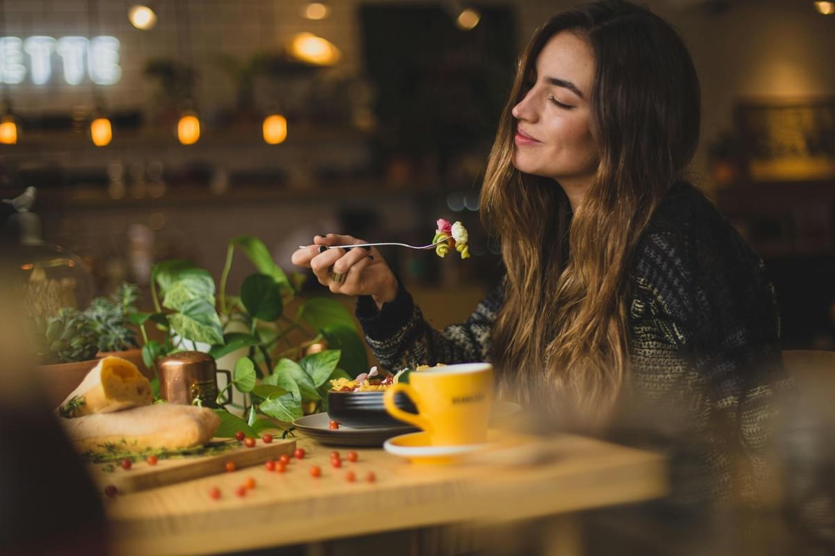 A woman sits at a wooden table in a warmly lit cafe, eyes closed as she prepares to enjoy a bite of colorful pasta. The scene is cozy and inviting, featuring fresh bread, lush greenery, and a bright yellow coffee cup in the foreground.