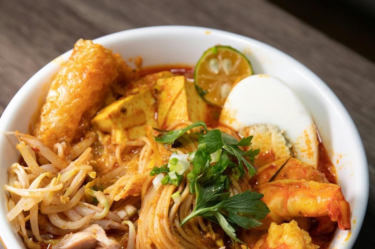 A bowl of Laksa featuring noodles topped with meat, eggs, and colorful vegetables.