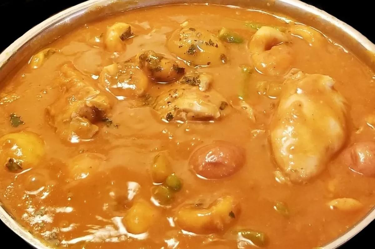 Close-up of a pot of thick, orange-red stew (possibly Manchamanteles Mole) with chicken pieces, potatoes, cherry tomatoes, and fruit (peaches or apricots) in a rich, savory sauce. 