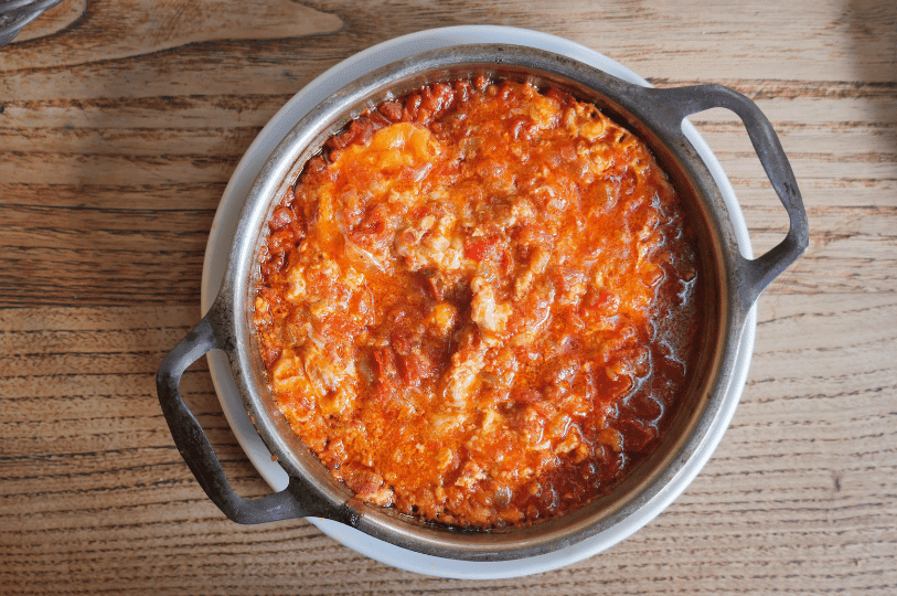 A rustic black pot filled with Turkish menemen—softly scrambled eggs nestled in a vibrant, spiced tomato stew—sits atop a white plate on a wooden surface, radiating warmth and homestyle comfort.