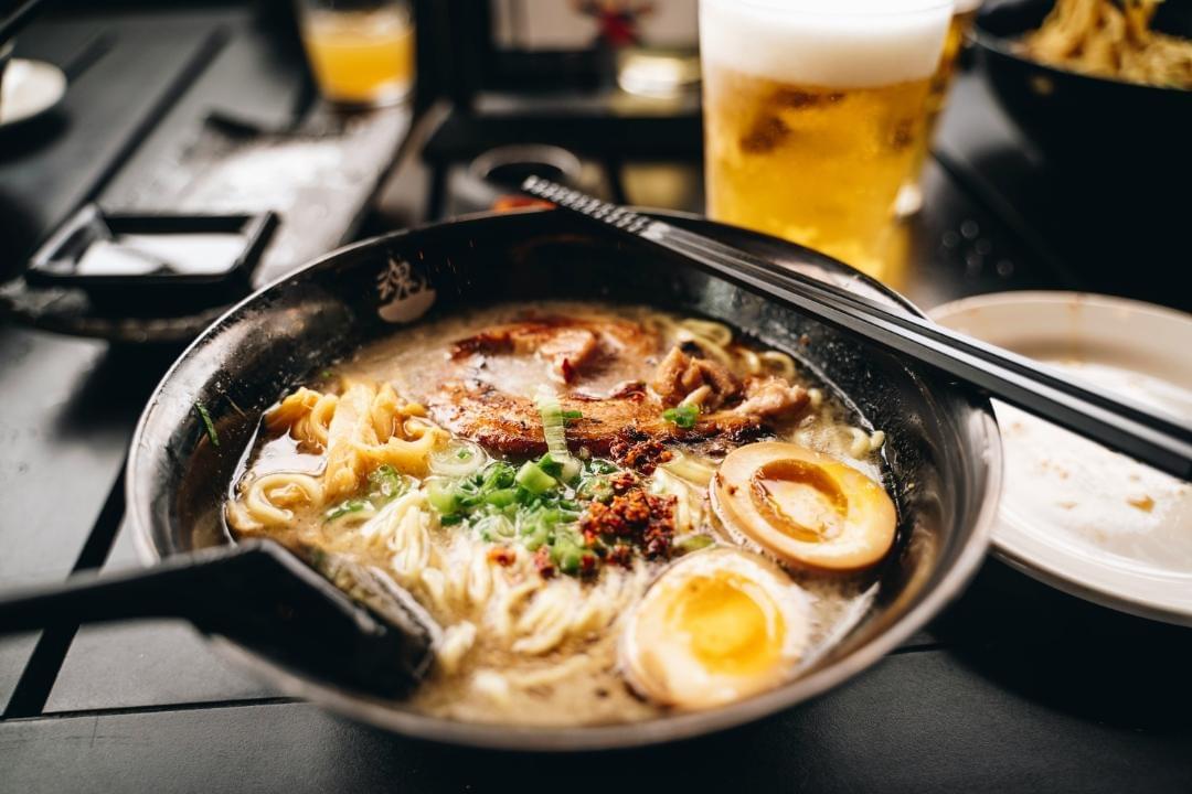 A steaming bowl of ramen, topped with soft-boiled eggs, chashu pork, and chopped green onions. A pair of chopsticks rests across the rim of the black bowl, while a tall glass of frothy beer stands softly blurred in the background.