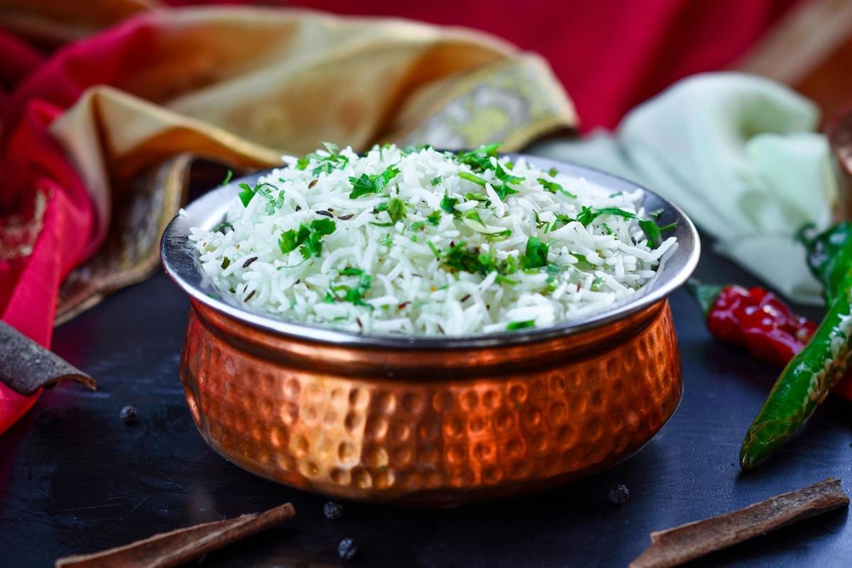 A hammered copper bowl overflows with fluffy white rice garnished with fresh green herbs and cumin seeds. The dish is set on a dark surface surrounded by scattered spices, including cinnamon sticks and chilies, with colorful fabric in the background.