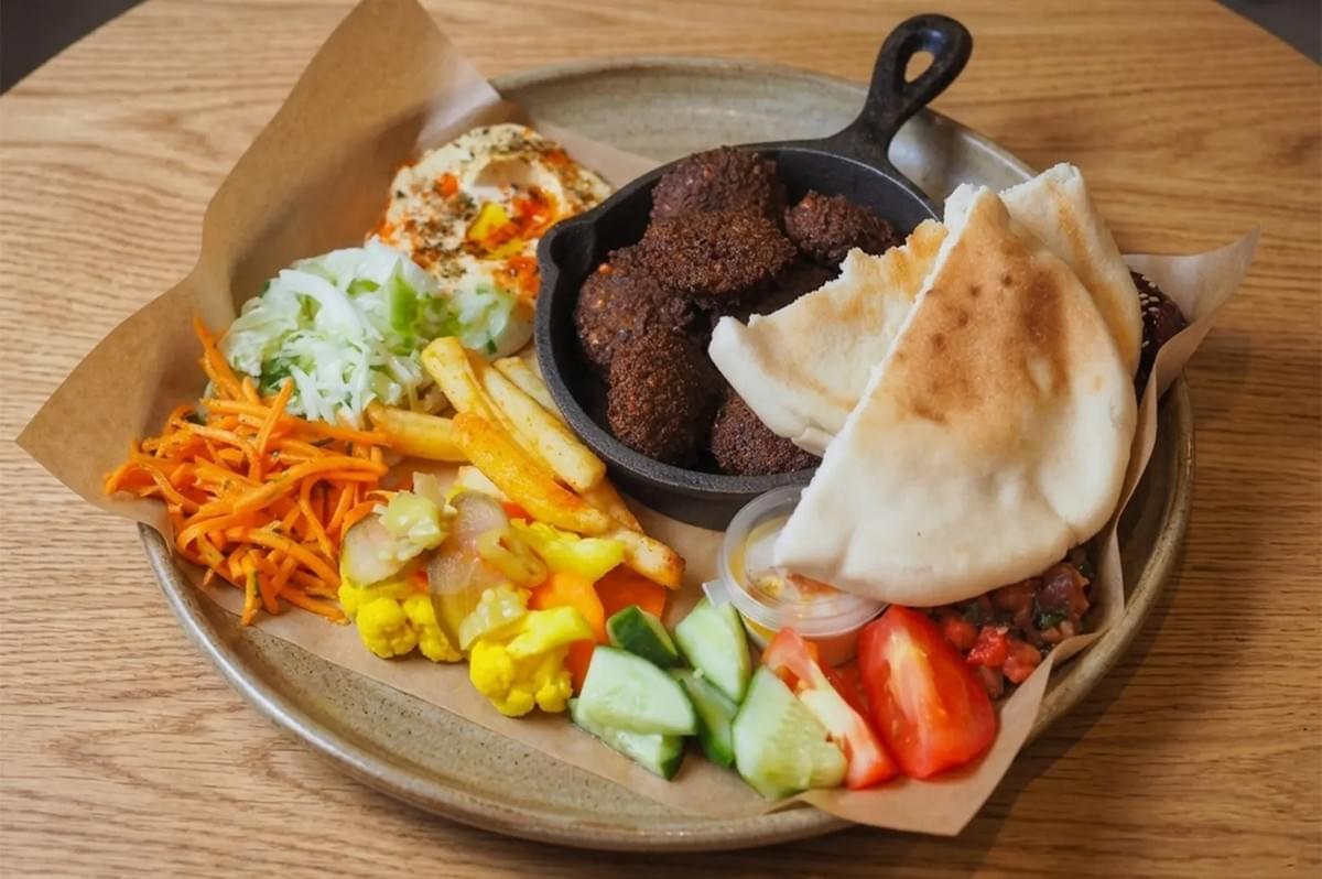 Middle Eastern falafel platter with pita bread, hummus, fresh vegetables, pickles, and fries on a wooden table.