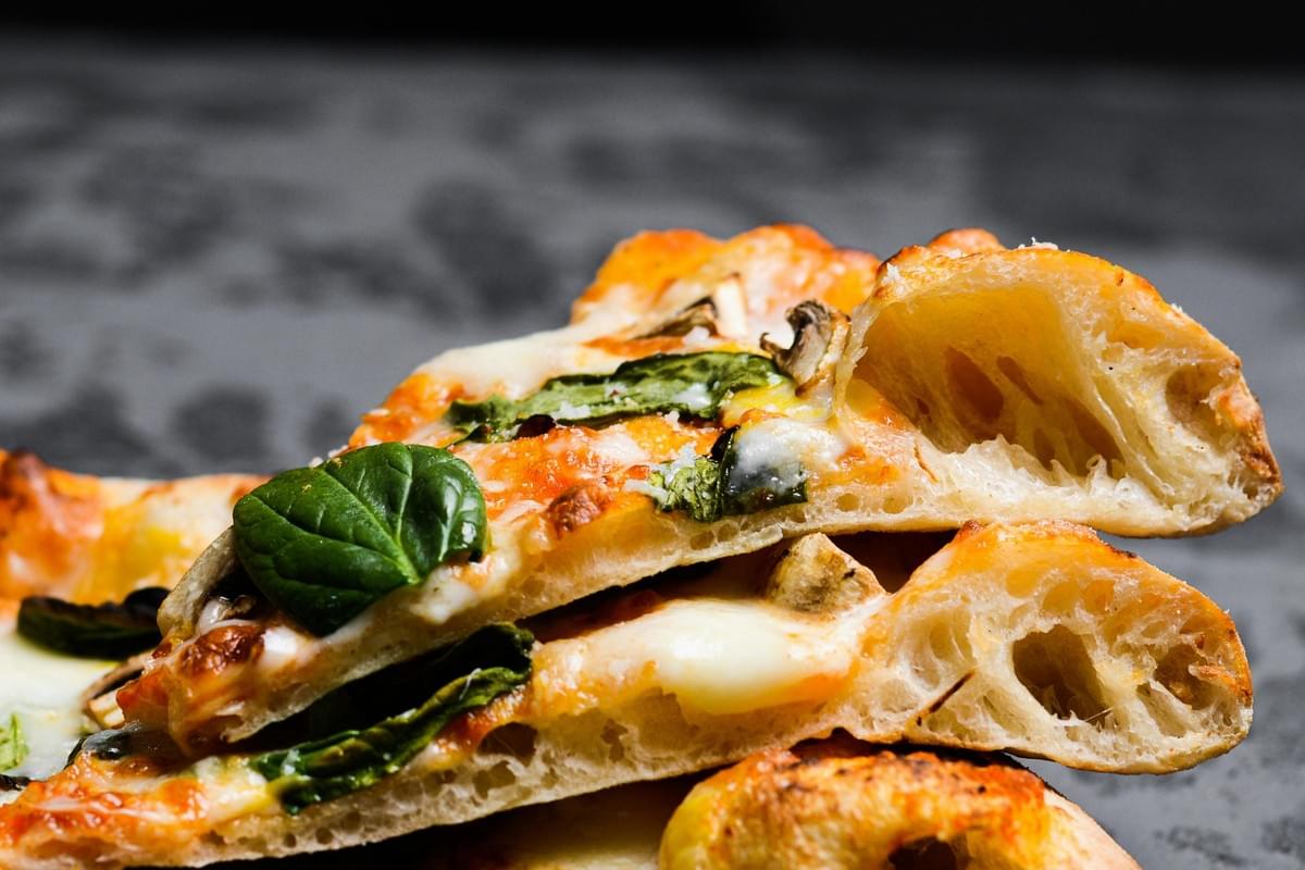 A close-up shot shows slices of freshly baked pizza stacked on top of each other against a dark, textured background. The focus highlights a thick, airy crust with large air pockets, topped with melted mozzarella cheese, mushrooms, and fresh green basil leaves.