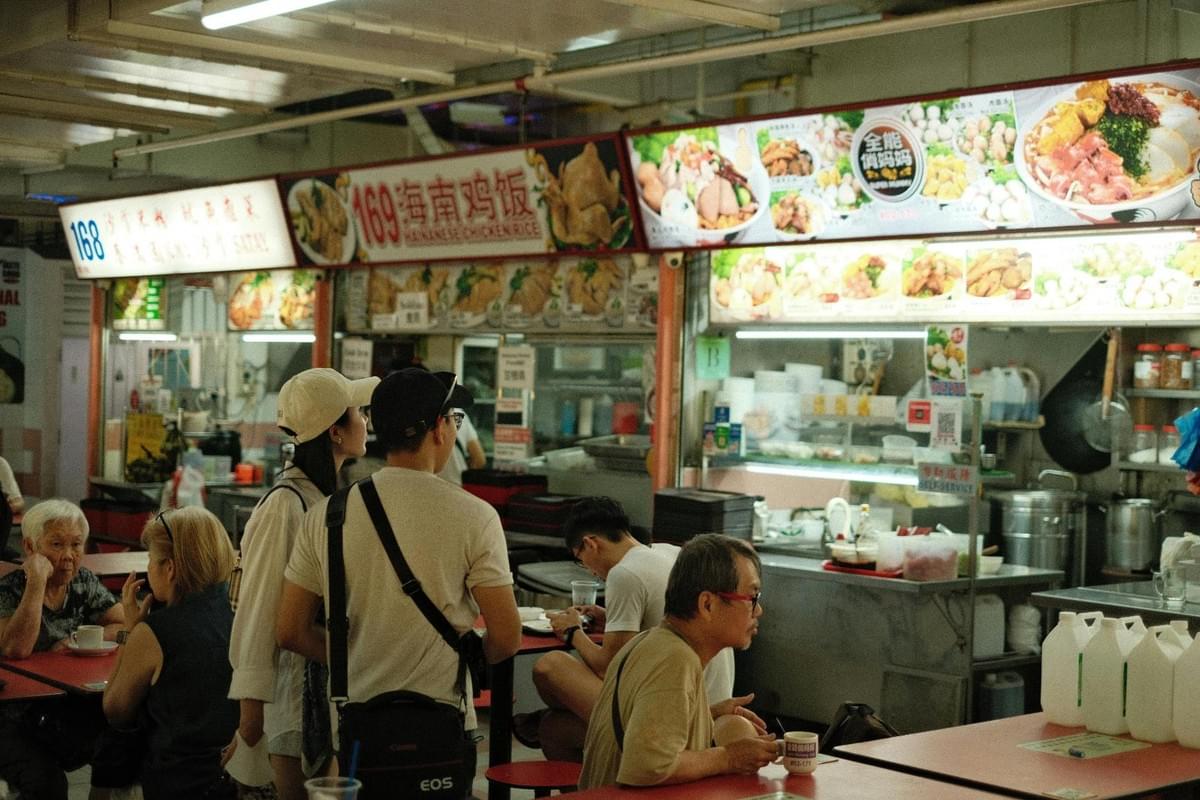 Customers gather at a bustling indoor hawker center featuring various food stalls with colorful overhead menus. People are seen seated at red tables or standing while they wait for their meals in the lively, casual atmosphere.