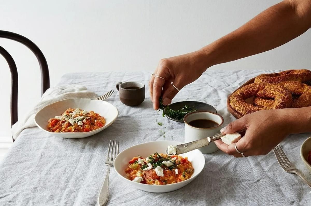 Hands garnishing bowls of Menemen (Turkish scrambled eggs) with herbs, served with coffee and a basket of Simit bread rings