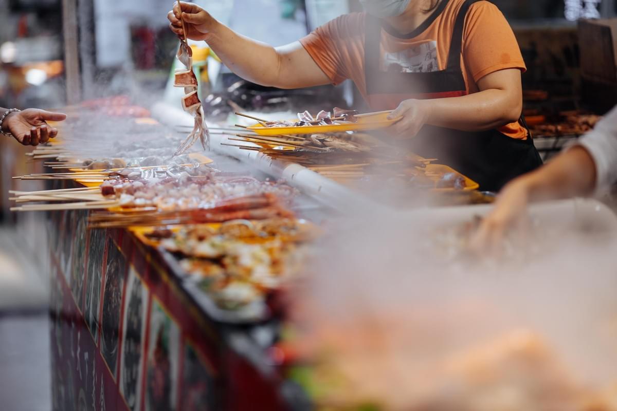 A vendor at a bustling Asian market prepares various skewers of fresh seafood, holding up a whole squid for a customer over a steaming grill display. Rows of diverse, uncooked skewered meats and seafood line the colorful stall counter amidst a haze of rising smoke.