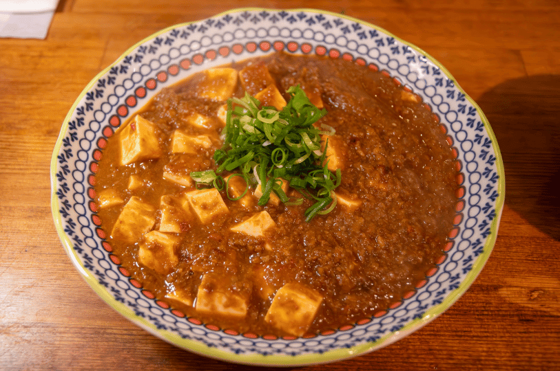 A bowl of spicy Mapo Tofu featuring a rich, thick gravy and plenty of minced meat, garnished with a generous pile of sliced scallions in a patterned blue and white bowl.