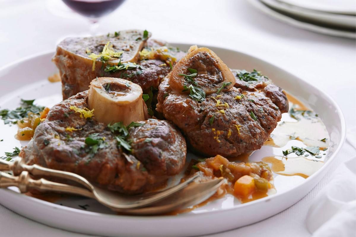 Three pieces of Osso Buco plated on a wide white dish, garnished with Gremolata (chopped parsley, lemon zest, garlic), and served with the vegetable braising sauce.