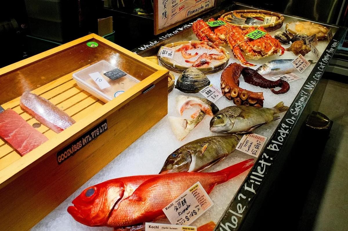 A seafood display case features a variety of fresh items laid out on a bed of crushed ice, including a bright red fish, whole fish, octopus tentacles, shrimp, and large crabs. Beside the ice bed, a wooden counter displays neatly sliced cuts of raw fish ready for preparation.