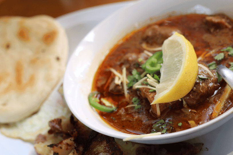 A close-up shot of a rich beef Nihari topped with a fresh lemon wedge and green chilies, served in a white bowl alongside a piece of fluffy flatbread.