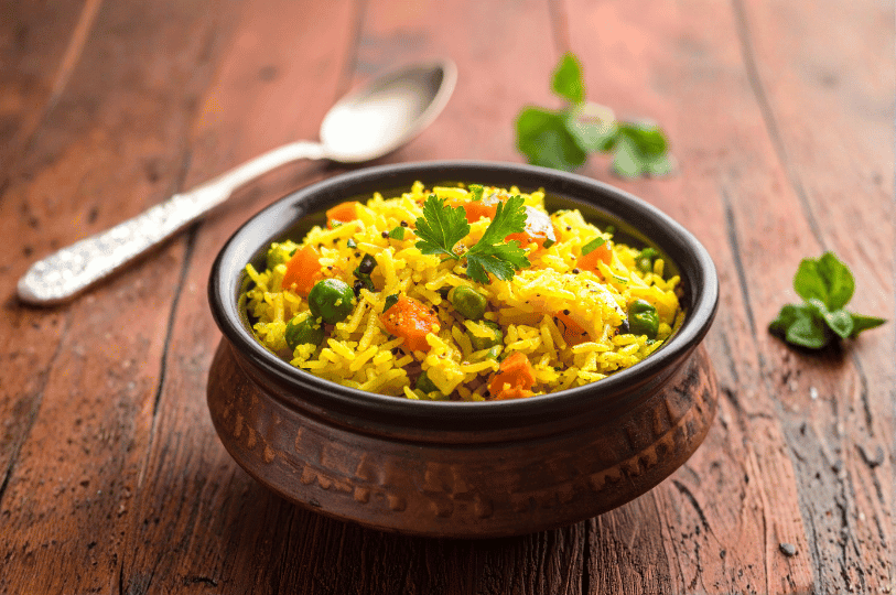A small, dark rustic bowl of yellow rice cooked with diced carrots and green peas, placed on a textured wooden table next to a vintage silver spoon.
