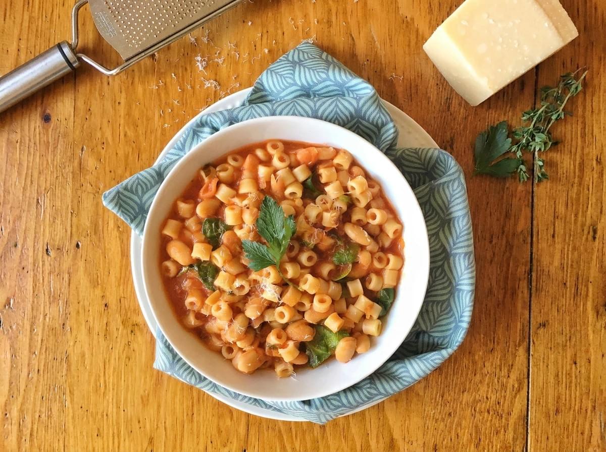 A bowl of ditalini pasta and bean soup topped with fresh herbs, resting on a blue cloth next to a grater and block of cheese.