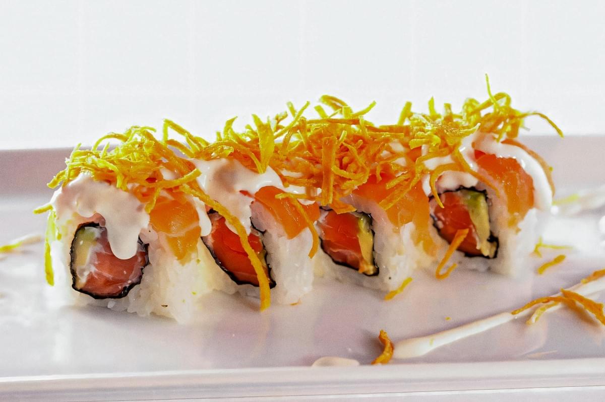 Creamy-topped salmon and avocado maki roll on a white plate, garnished with crispy tempura flakes and drizzled sauce for a gourmet sushi presentation.