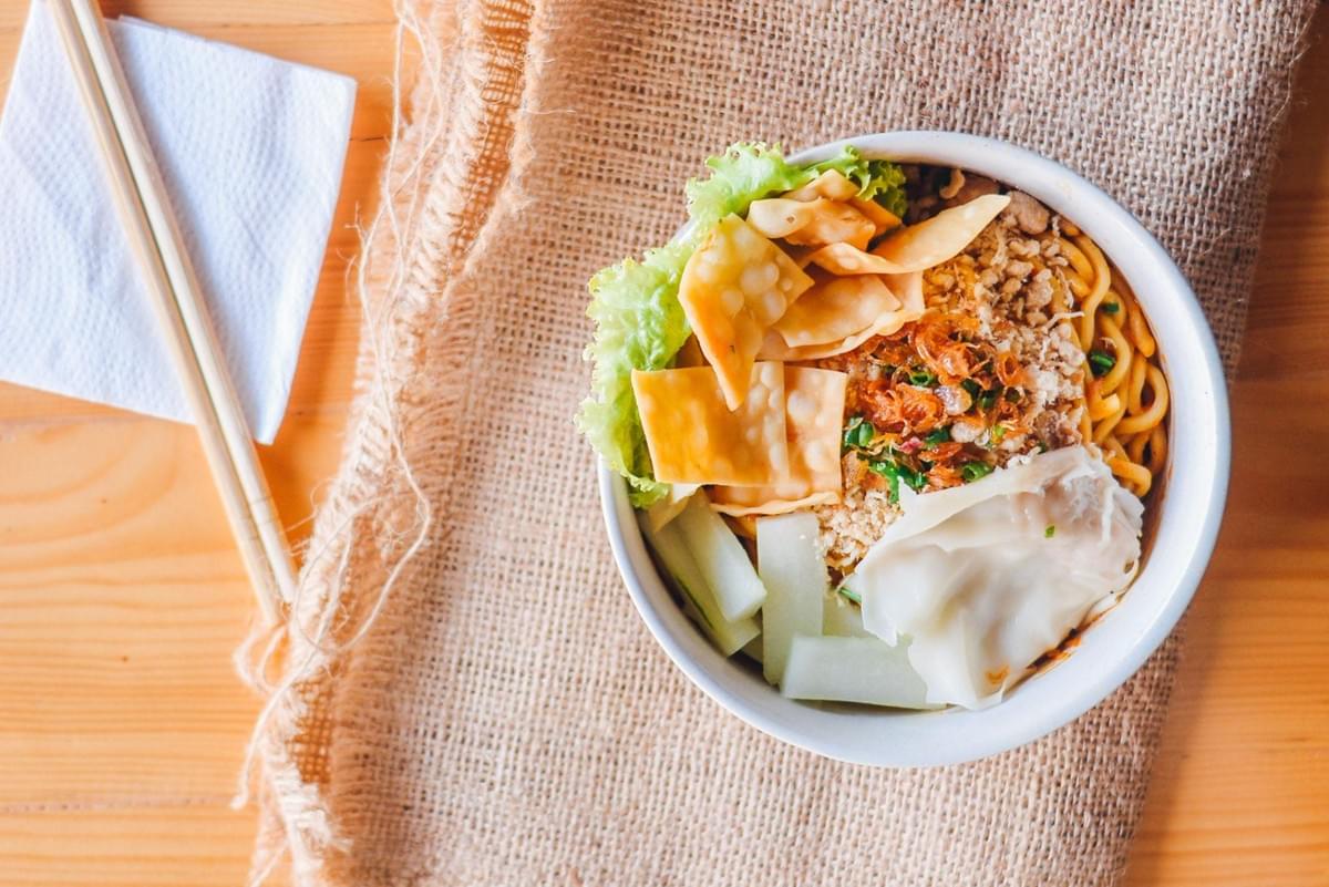 A top-down view shows a white bowl filled with noodles, fried wonton crisps, steamed wontons, lettuce, sliced cucumbers, and minced meat toppings. The bowl rests on a burlap cloth next to wooden chopsticks resting on a white napkin.