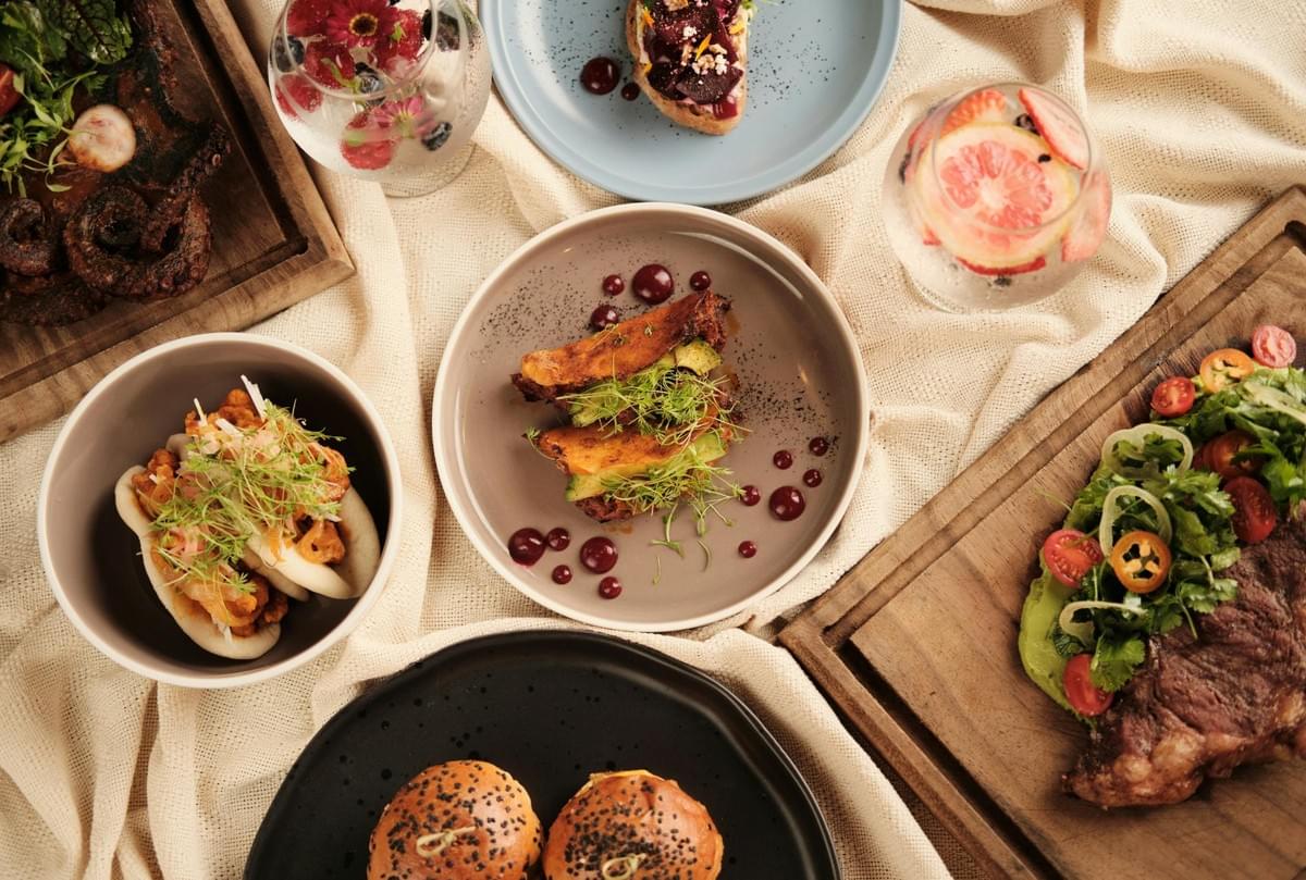 An overhead shot showcases a vibrant spread of gourmet dishes and fruity cocktails arranged on a light beige cloth. The feast includes bao buns, avocado tacos, sliders, grilled meats, and fresh salads served on colorful plates and wooden boards.