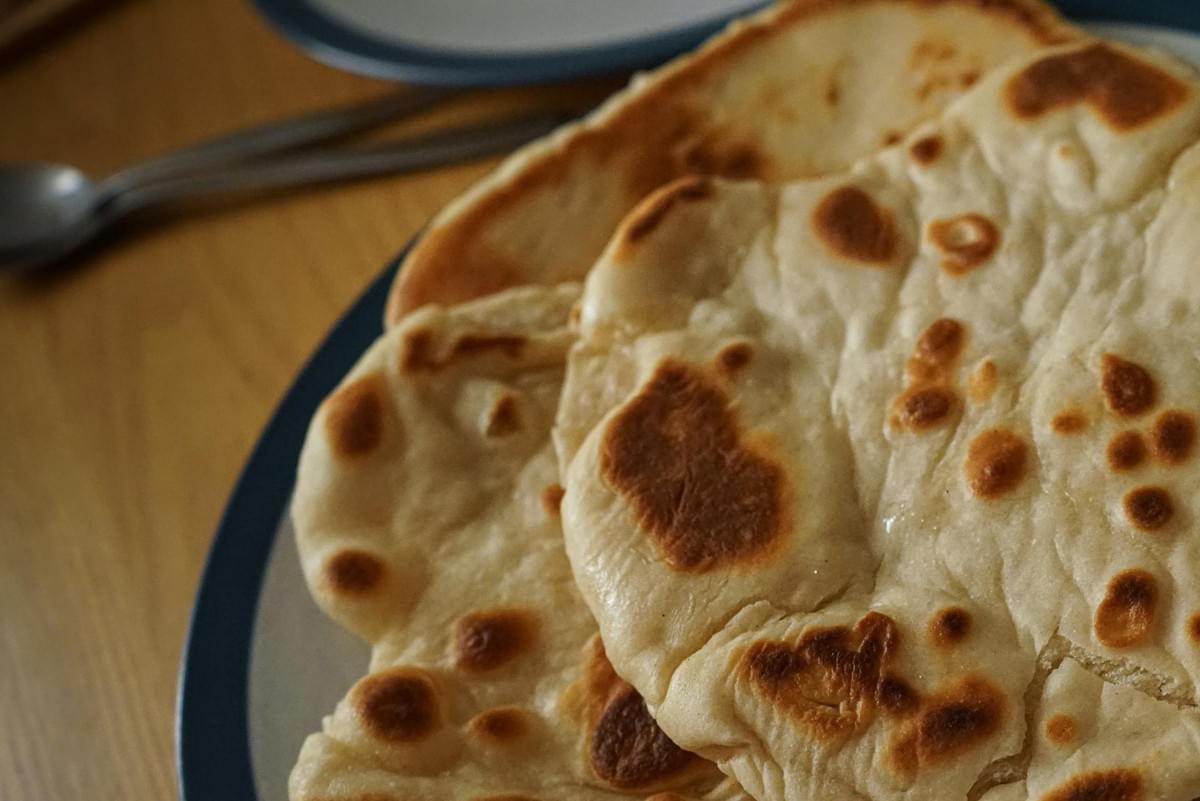 A stack of warm, golden-brown flatbreads with charred spots rests on a blue-rimmed plate. In the background, a wooden table holds cutlery and another dish out of focus.