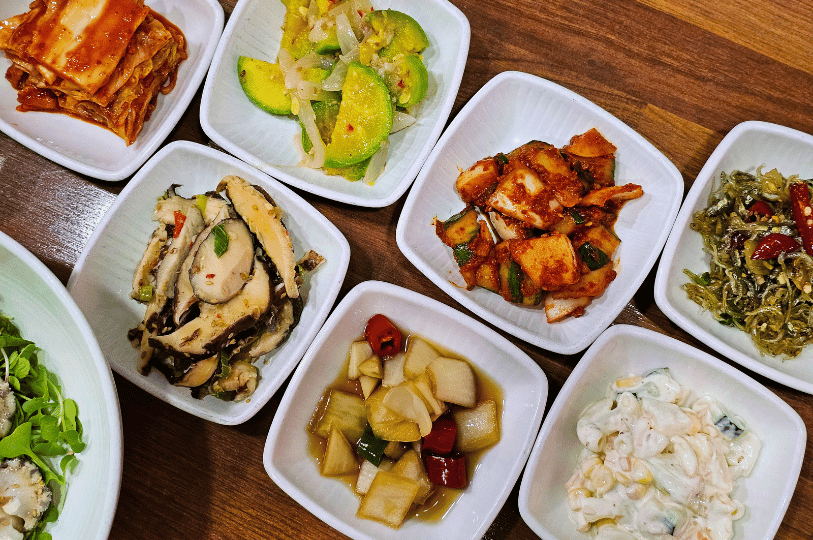 A tight, colorful arrangement of eight square white bowls on a wooden surface, showcasing a diverse selection of Korean banchan such as cubed radish kimchi, sautéed mushrooms, stir-fried zucchini, pickled onions, and macaroni salad.