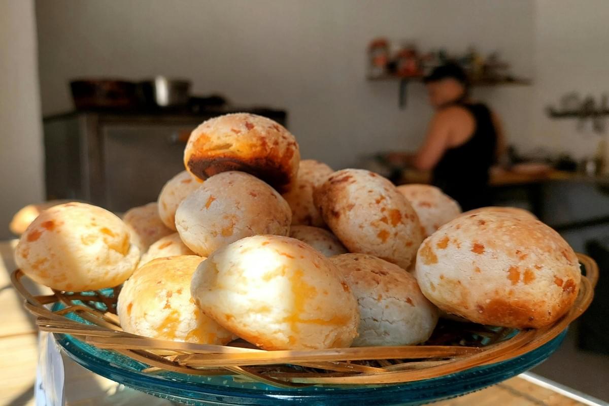A mound of golden, round cheese breads rests in a shallow basket, glowing warmly under bright, directional sunlight. In the soft-focus background, a person works in a kitchen setting, suggesting the pastries are freshly baked.