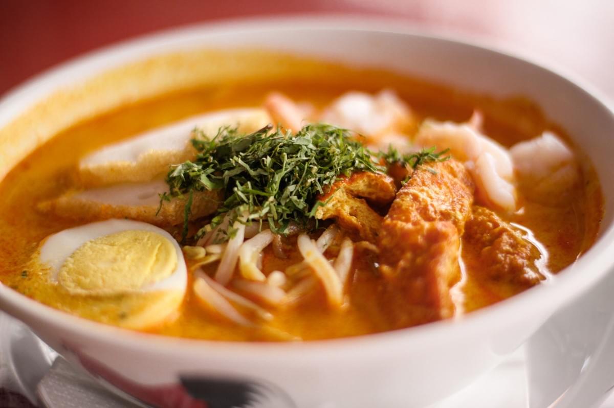 A bowl of Laksa soup filled with meat and colorful vegetables, garnished with herbs.