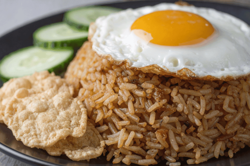 A macro shot focusing on the texture of seasoned INasi Goreng, crowned with a crispy-edged fried egg and accompanied by golden kerupuk and fresh cucumber rounds.
