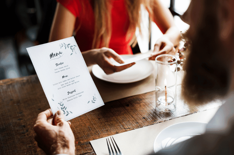 A couple dining at a rustic wooden restaurant table; the focus is over the shoulder of a person holding a minimalist menu listing "Starter," "Main," and "Dessert" options while sitting across from a woman in a red dress.