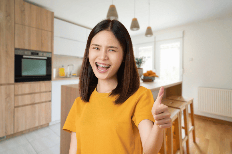 A smiling woman wearing a yellow t-shirt giving a thumbs-up in a modern, well-lit kitchen.