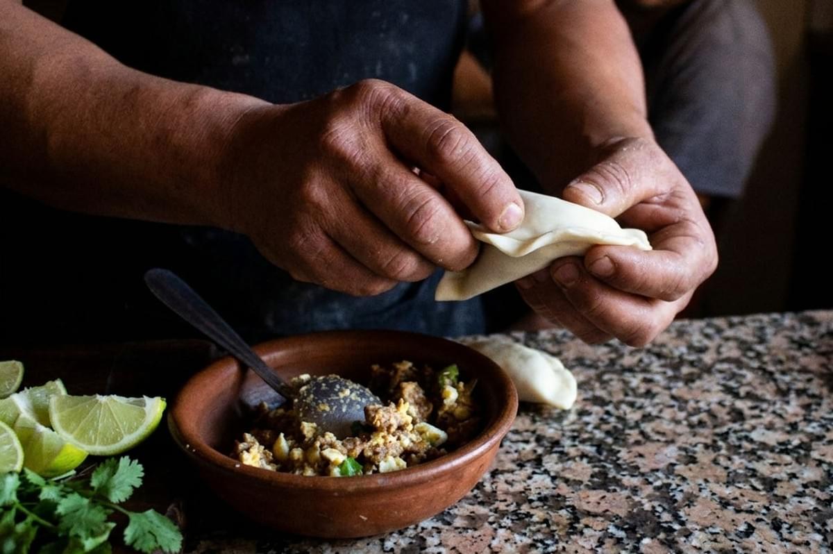 Hands pinching and folding empanada dough using traditional repulgue braiding technique with filling bowl and fresh ingredients nearby