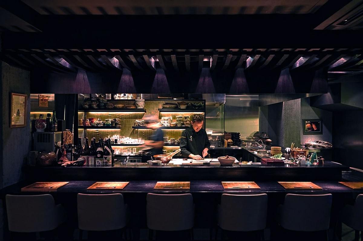 A dimly lit restaurant kitchen with a chef focused on preparing food. The countertop is neat, bottles and utensils are visible, creating an intimate and professional ambiance.