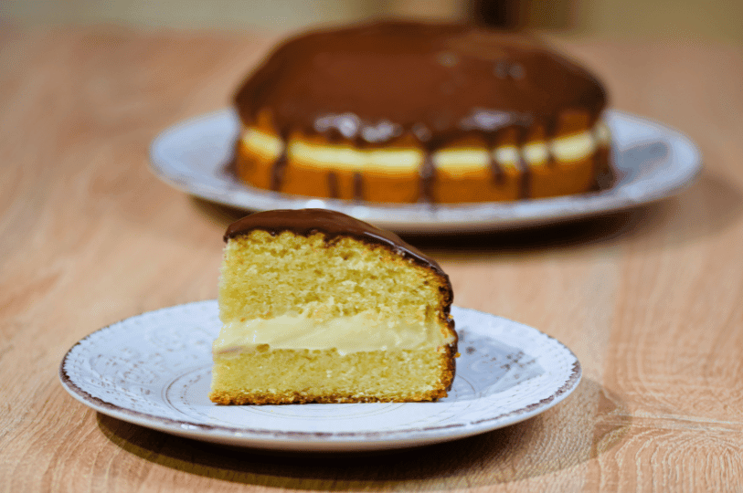 A close-up of a single slice of Boston Cream Pie on a white decorative plate, highlighting the even ratio of airy sponge cake to creamy custard filling, with the whole cake blurred in the background.