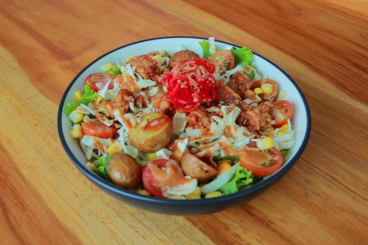  This vibrant bowl of salad features a mix of fresh greens, cherry tomatoes, corn, and roasted potatoes topped with crispy protein pieces and a drizzle of creamy dressing. A bright nest of shredded red cabbage or pickled vegetables sits at the center, all presented in a dark bowl on a warm wooden surface.
