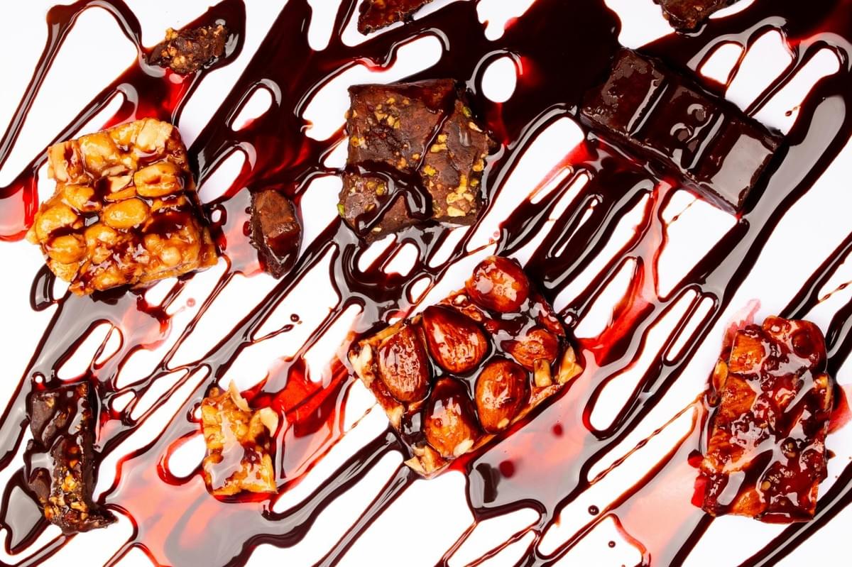 An assortment of nut clusters, brownies, and chocolate pieces is arranged on a white surface and heavily drizzled with dark chocolate and reddish syrup. The overhead shot captures the glossy textures of the sweets and the messy, artistic swirls of the sauces.