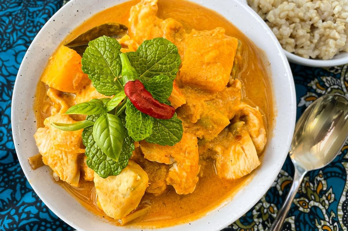 A bowl of authentic Thai Massaman chicken curry with chunks of potato and pumpkin, garnished with fresh mint, basil, and a red chili, served with a side of brown rice.