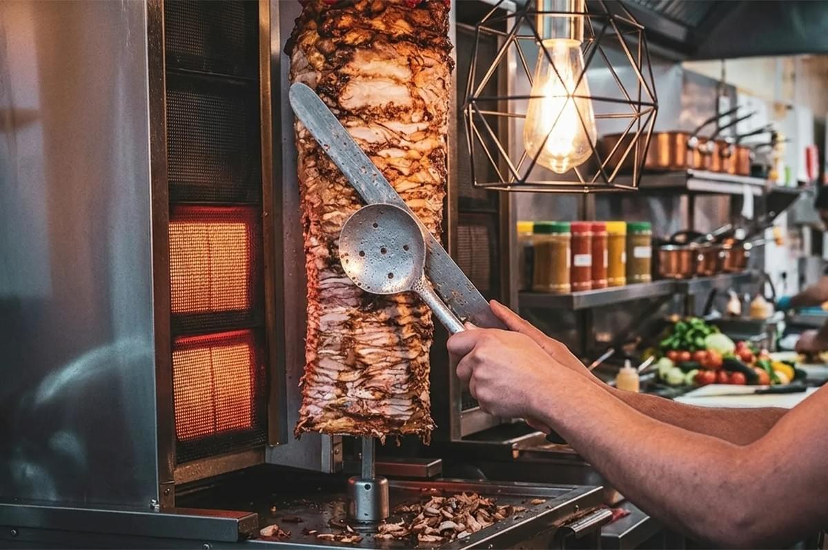 Close-up of a chef slicing seasoned meat from a vertical rotisserie grill for shawarma or doner kebab in a restaurant kitchen.