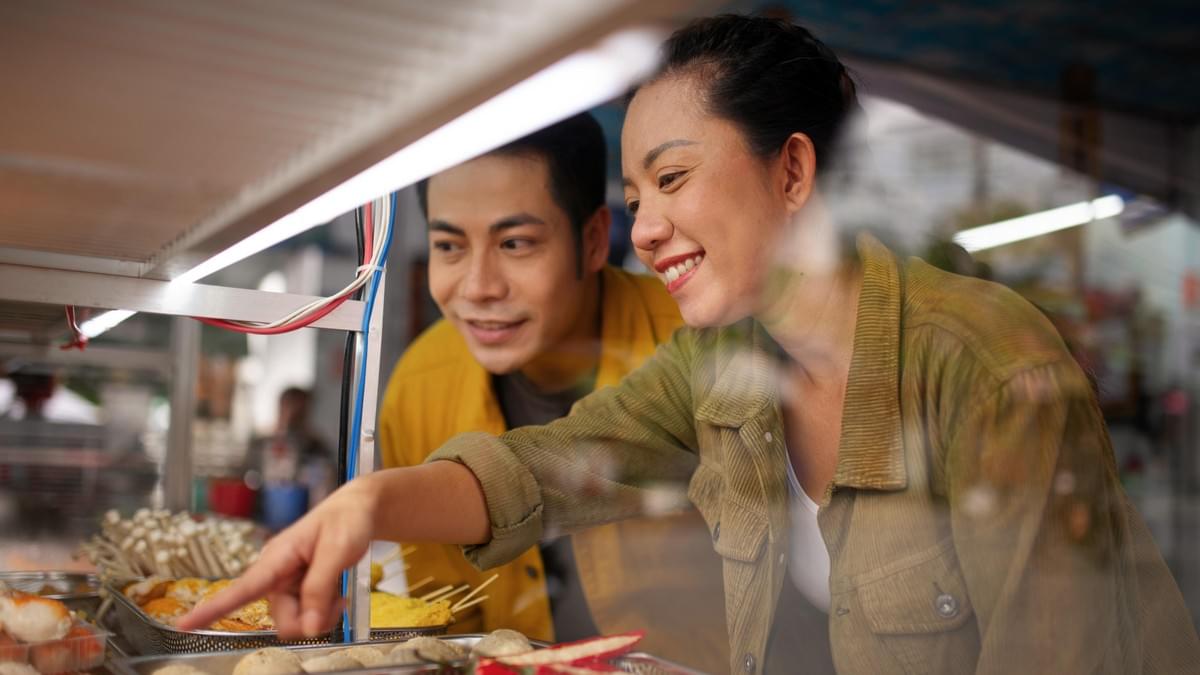  This medium shot captures a smiling woman pointing to street food displayed behind a glass counter, with a man looking on just behind her.